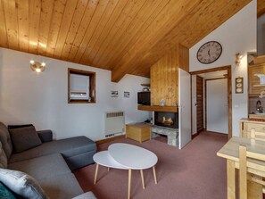A cozy room with a wooden ceiling, a fireplace, a sofa, a small table, and a kitchen area with wooden cabinets. - Apartment 5 guests, clear view, at the slopes (Val-d'Isère)