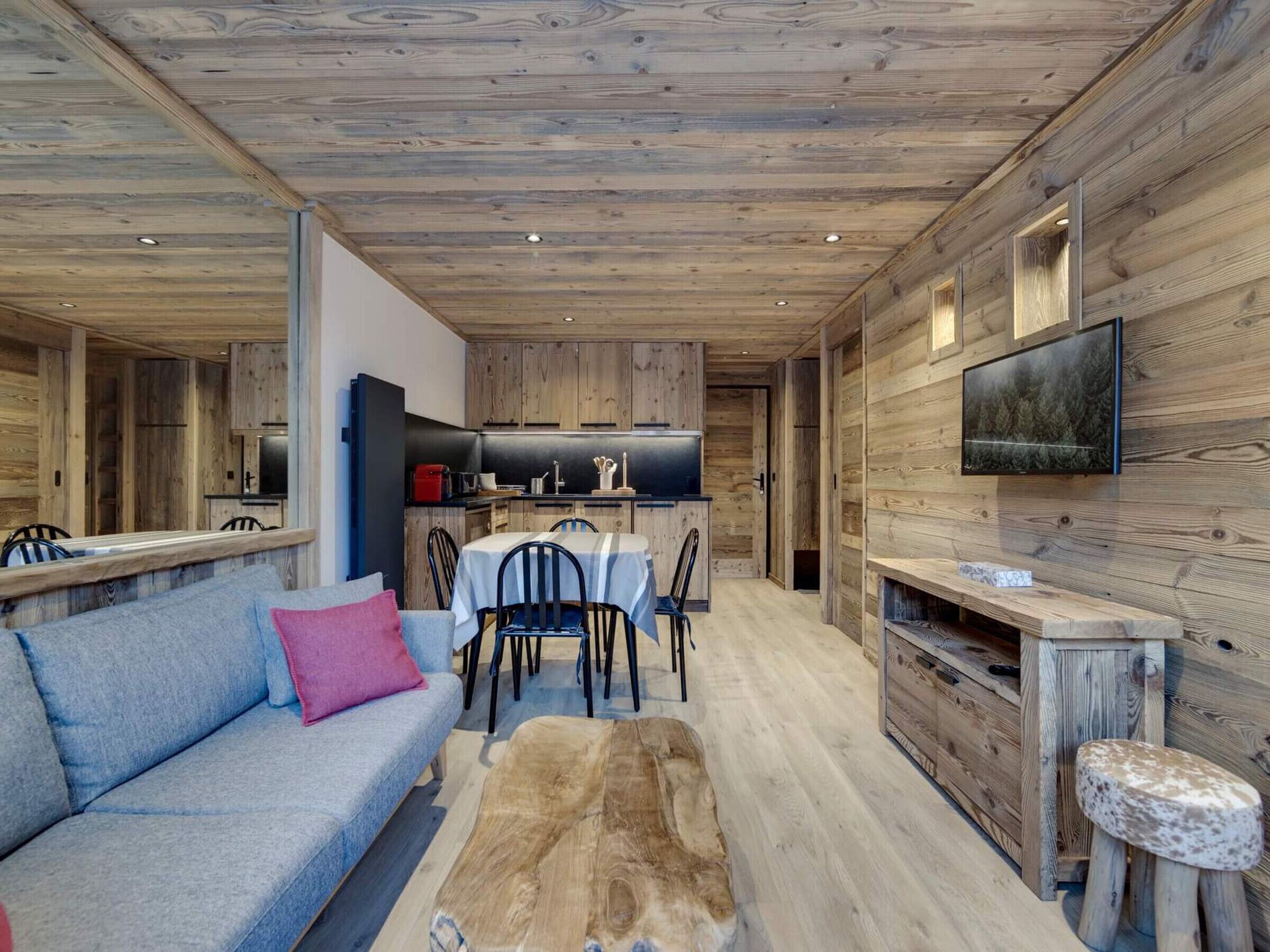 A modern interior with a wooden ceiling, a dining area with a table and chairs, a sofa with a pink pillow, and a TV mounted on the wall.