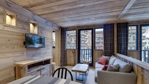 Ein gemütliches Blockhaus-Interieur mit Holzbalkendecke, einem an der Wand montierten Fernseher, einer Couch, einem Esstisch und Blick auf einen Balkon mit Gebäuden.