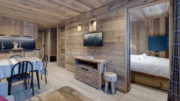 Ein modernes, rustikales Zimmer mit Holzbalkendecke, einem Essbereich mit Tisch und Stühlen, einem an der Wand befestigten Fernseher und einem Bett mit Blick durch eine Glastür.