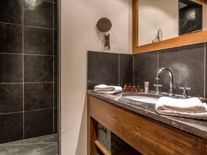 Un bagno moderno con una parete rivestita in piastrelle scure, un mobile lavabo in legno, uno specchio rotondo, un lavandino con rubinetto cromato e asciugamani ordinatamente piegati.