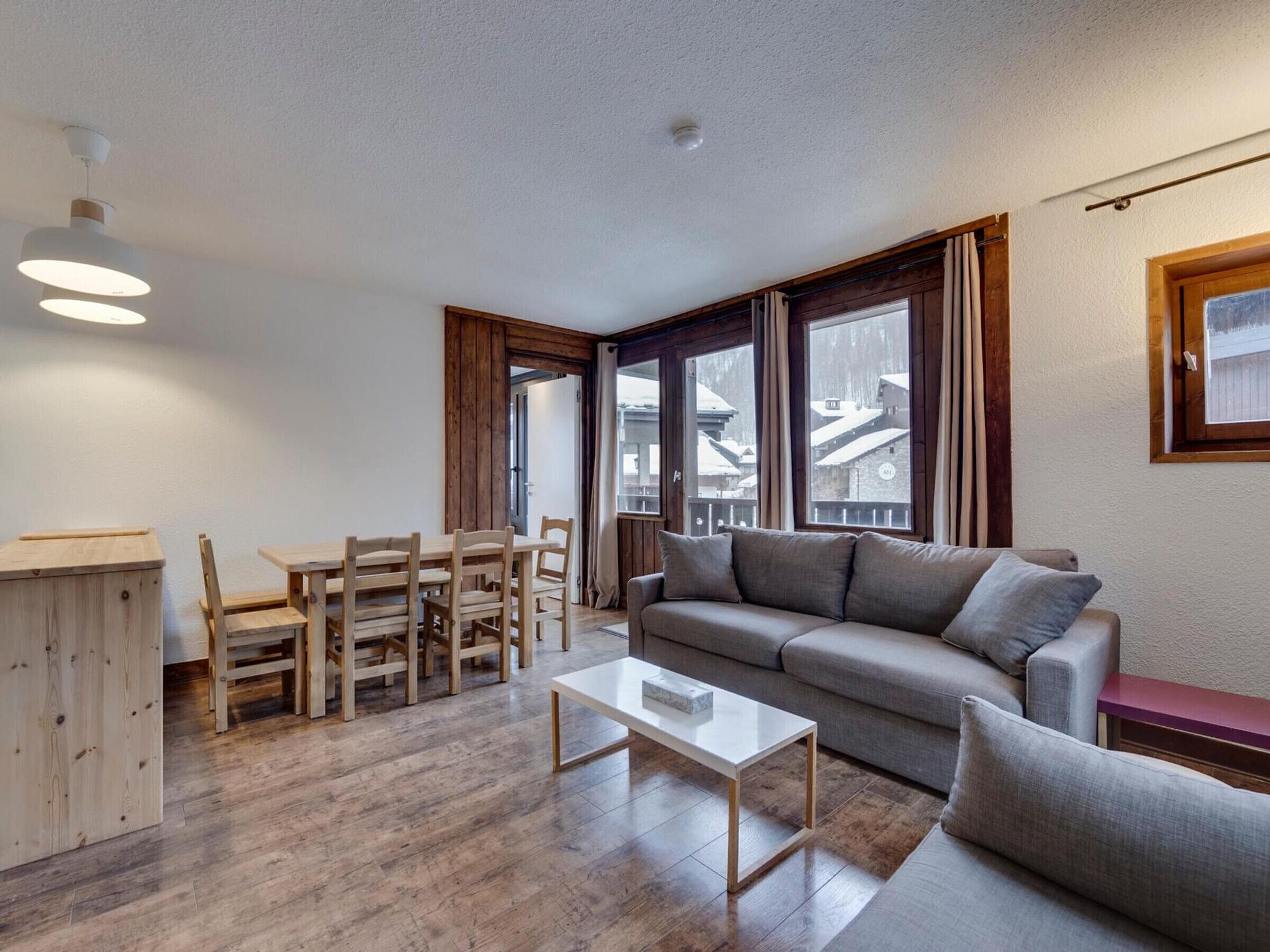 Ein modernes Wohnzimmer mit einem Holzes Esstisch, einem grauen Sofa und einem weißen Couchtisch.