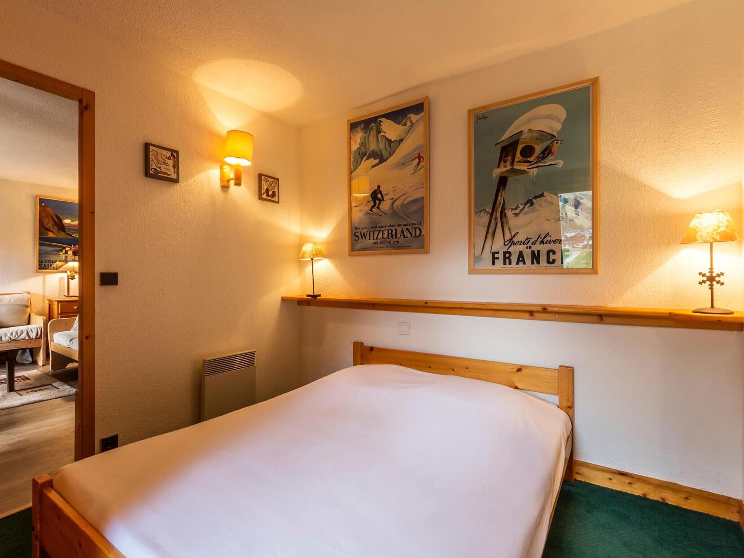 A hotel room with a bed, two wall-mounted lamps, a wooden headboard, and two framed posters advertising Switzerland and France.