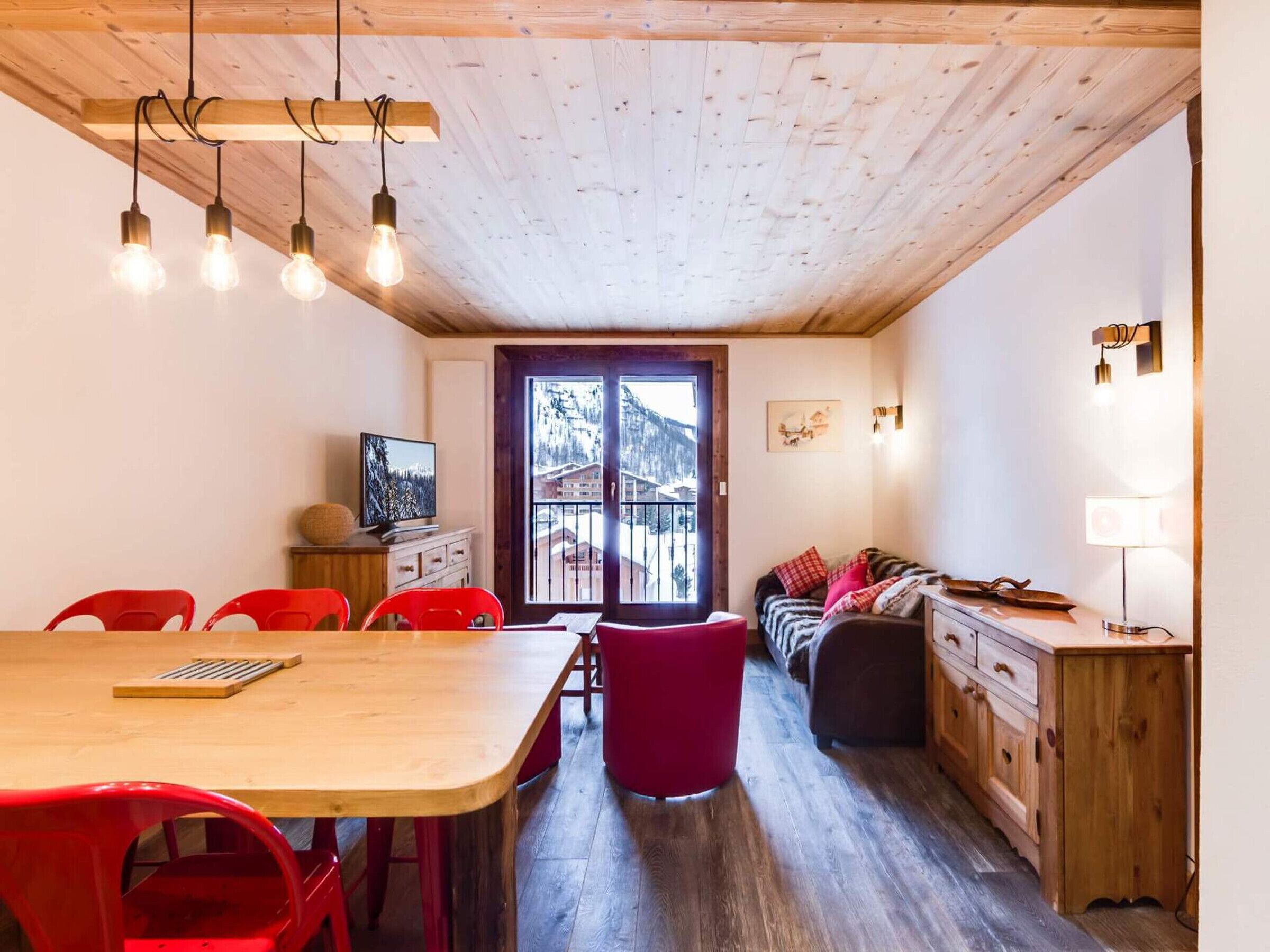 A cozy room with a wooden ceiling, a dining table with red chairs, a sofa, and a TV on a wooden cabinet.
