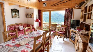 A cozy cabin interior with a dining table, chairs, a sofa, a television, and a view of the mountains. - Charming apartment - 6 guests Saint-Jean-de-Sixt (Saint-Jean-de-Sixt)
