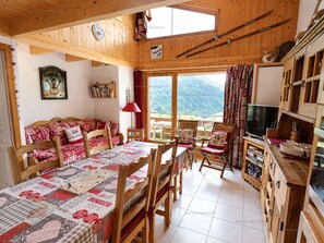 Ein gemütliches Blockhäuschen-Interieur mit Esstisch, Stühlen, einer Couch, einem Fernseher und Blick auf die Berge.