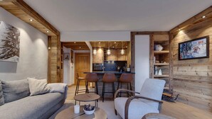 Een moderne woonkamer met een bank, fauteuils en een salontafel. In de achtergrond is een keuken zichtbaar met barstoelen.