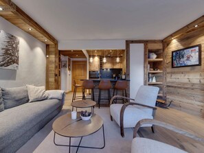 A modern living room with a sofa, armchairs, and a coffee table. A kitchen is visible in the background with bar stools. - Cosy apartment at the slopes - 4 guests (Val-d'Isère)