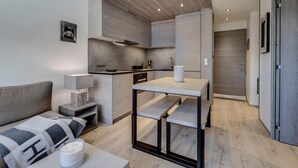 Une cuisine moderne dotée d’un four encastré, une table à manger avec des bancs, et un canapé gris dans l’espace salon.