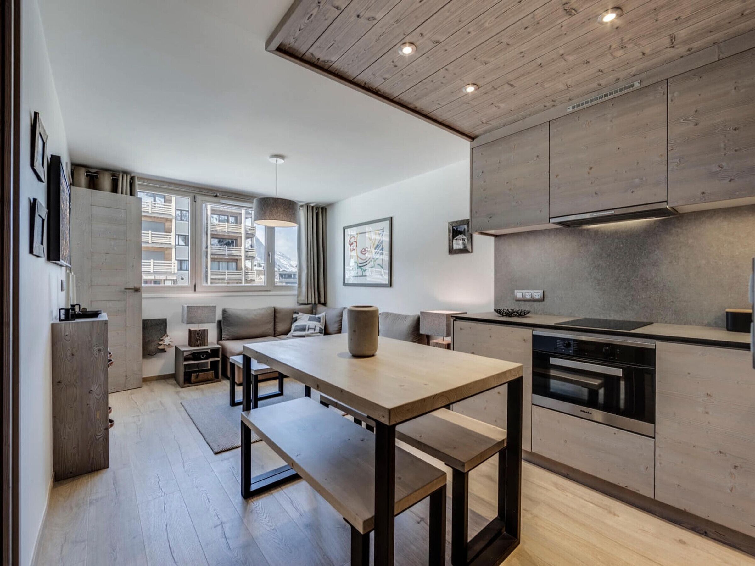 A modern kitchen with a built-in oven, a dining area with a wooden table and benches, and a view of a building through a window.