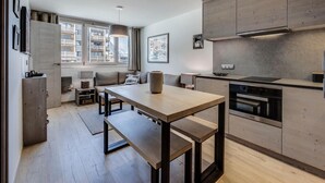 Une cuisine moderne dotée d’un four encastré, un coin repas avec une table et des bancs en bois, et une vue sur un immeuble par la fenêtre.