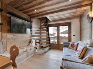 A cozy room with a wooden interior, a staircase, a sofa with patterned cushions, and a large window offering a view of a snowy landscape. - Spacious apartment near slopes - 4 guests (Val-d'Isère)