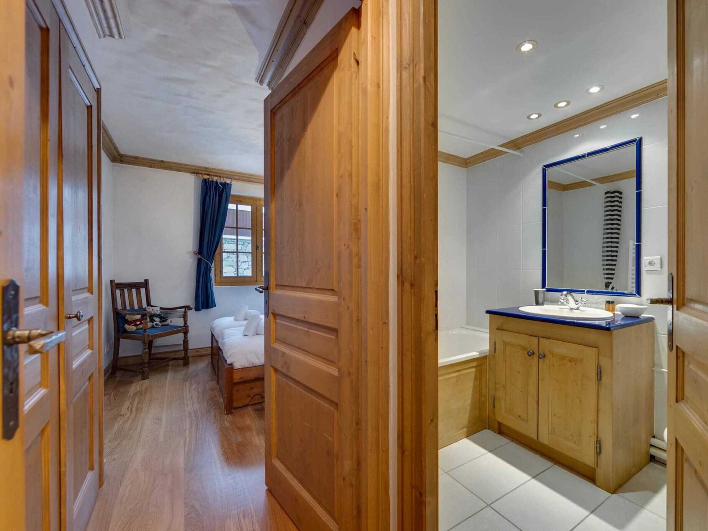 A hotel room with a wooden door, a bed, a chair with toys, a window with curtains, and a bathroom with a sink and mirror.