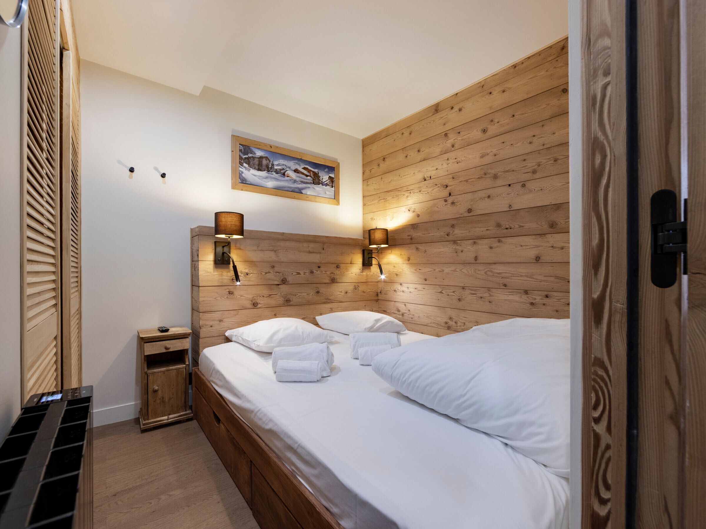 Een slaapkamer met een houten hoofdbord, twee bedlampen, een klein nachtkastje en een schilderij aan de muur.