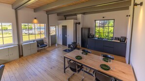 Ruime kamer met houten vloer, een eettafel gedekt met borden en glazen, een keuken met spoelbak en kastjes, en grote ramen met uitzicht op groen.