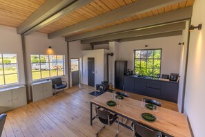 A spacious room with a wooden floor, a dining table set with plates and glasses, a kitchen area with a sink and cabinets, and large windows offering a view of greenery. - Warft workshop - Krummhörn (Krummhörn)