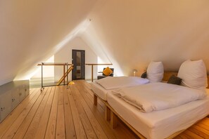 A room with a sloped ceiling, wooden flooring, a bed with white bedding, a desk with a chair, and a door. - Warft workshop - Krummhörn (Krummhörn)