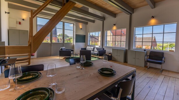 Een ruime kamer met een hoge plafond, grote ramen, een houten tafel gedekt met glazen en een trap.
