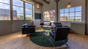 Een moderne woonkamer met een bank, fauteuils, een klein tafeltje en grote ramen met uitzicht op gebouwen en groen.