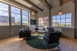 Ein modernes Wohnzimmer mit einer Couch, Sesseln, einem kleinen Tisch und großen Fenstern, die Blick auf Gebäude und Grünflächen bieten.