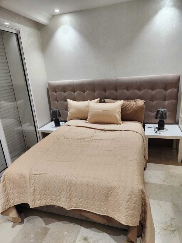 A bedroom with a large bed, two bedside tables with lamps, a sliding door, and a tiled floor.
