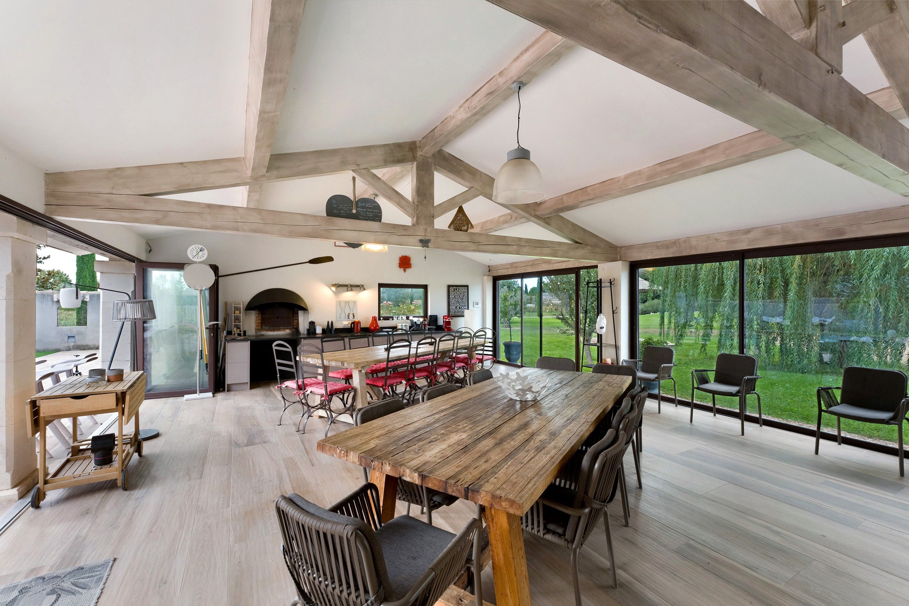 Un vaste espace à manger avec une grande table et des chaises en bois, une cuisine en arrière-plan et de grandes fenêtres donnant sur l’extérieur.