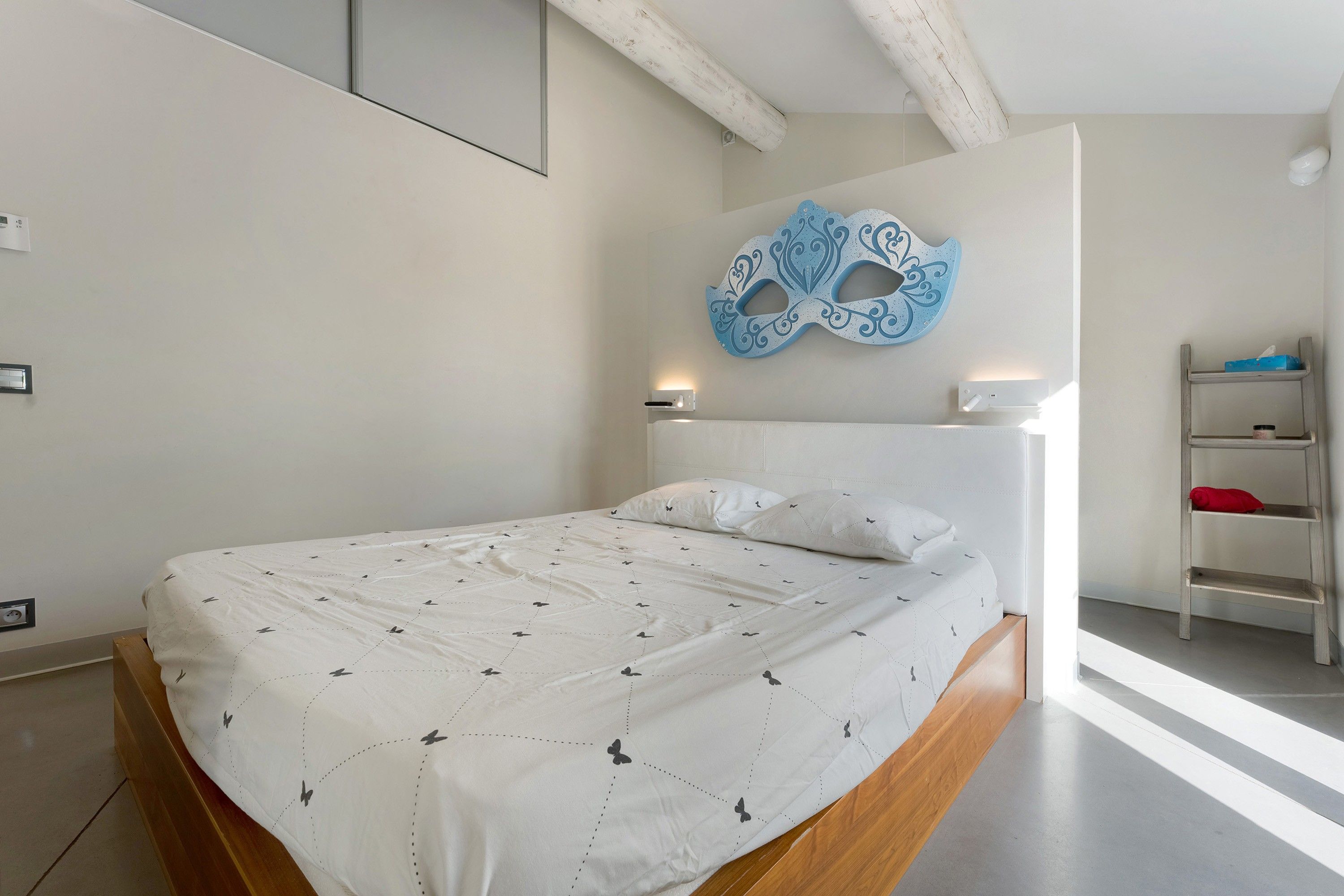 Une chambre à coucher moderne avec un lit, un masque décoratif accroché au mur et une petite étagère sur laquelle sont posés quelques objets.