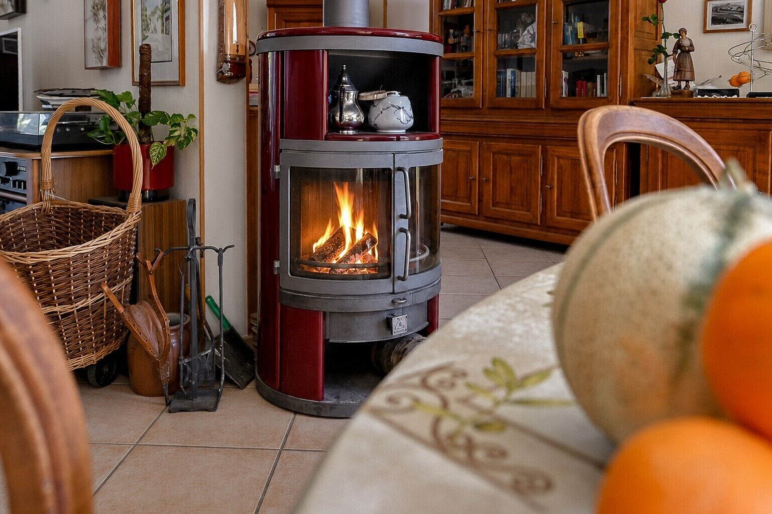 Una estufa de leña con puerta de vidrio, una cesta de mimbre y una silla en una habitación con armarios de madera y suelo de baldosas.