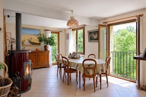 Un comedor con una mesa dispuesta para cuatro, una estufa de madera y un balcón con vistas a zonas verdes.