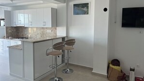 Une cuisine moderne avec des armoires blanches, un comptoir en marbre et une cuisinière à gaz. On y trouve deux tabourets de bar avec des sièges rembourrés. Un tableau est accroché au mur, et un téléviseur est fixé au mur.
