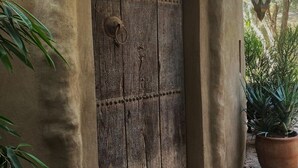Eine rustikale Holztür mit einem verzierten Metallring als Griff, eingebettet in einen pflastergelegten Eingangsbereich mit einem Blumentopf und einer Laterne.