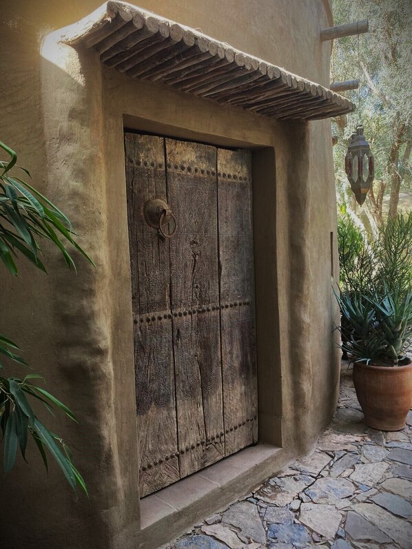 A rustic wooden door with a decorative metal ring handle, set in a stone-paved entrance area with a potted plant and a lantern.
