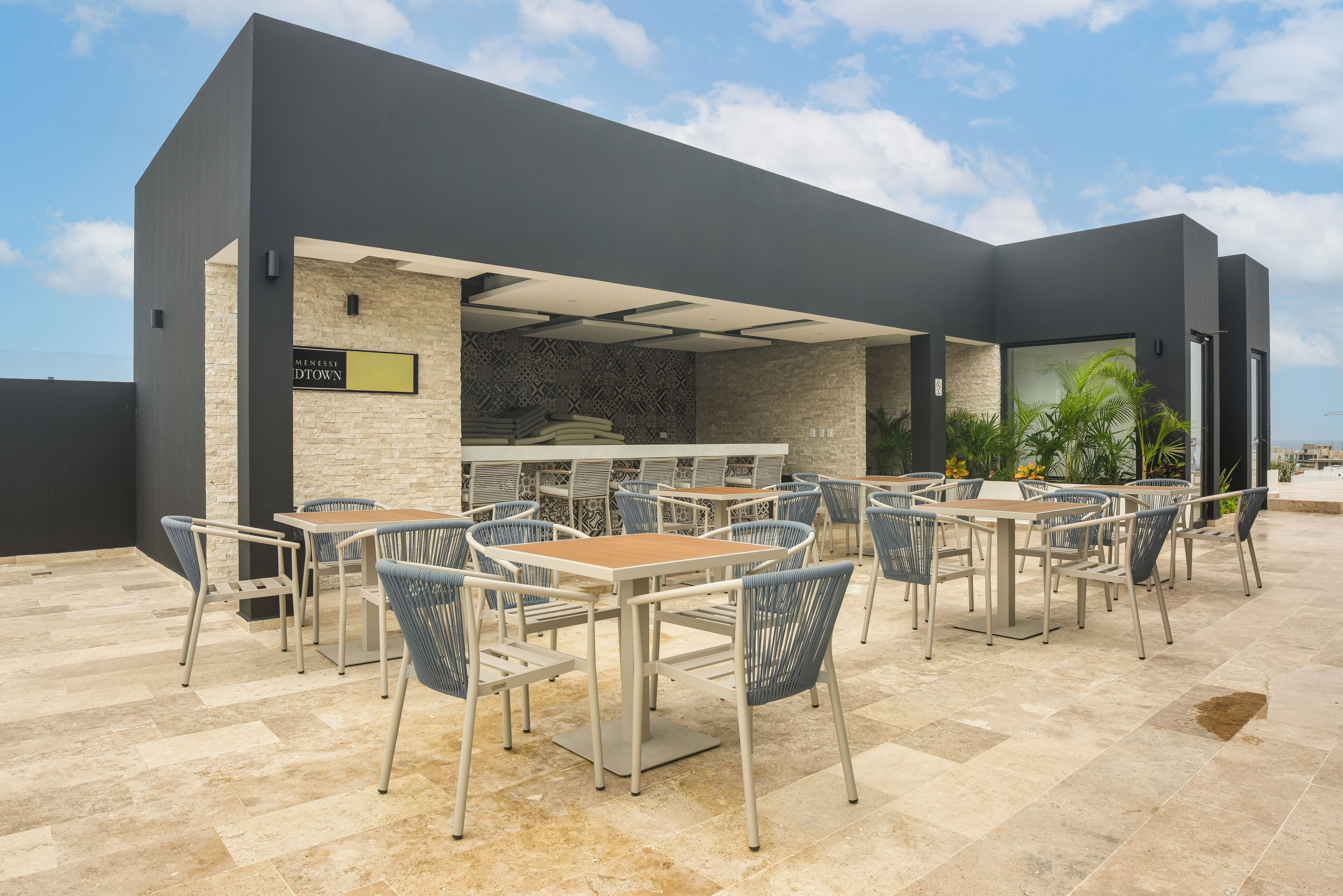 Un espace extérieur aménagé pour les repas, avec des tables et des chaises, un mur en pierre et une enseigne indiquant « MENGES DTDOWN ».