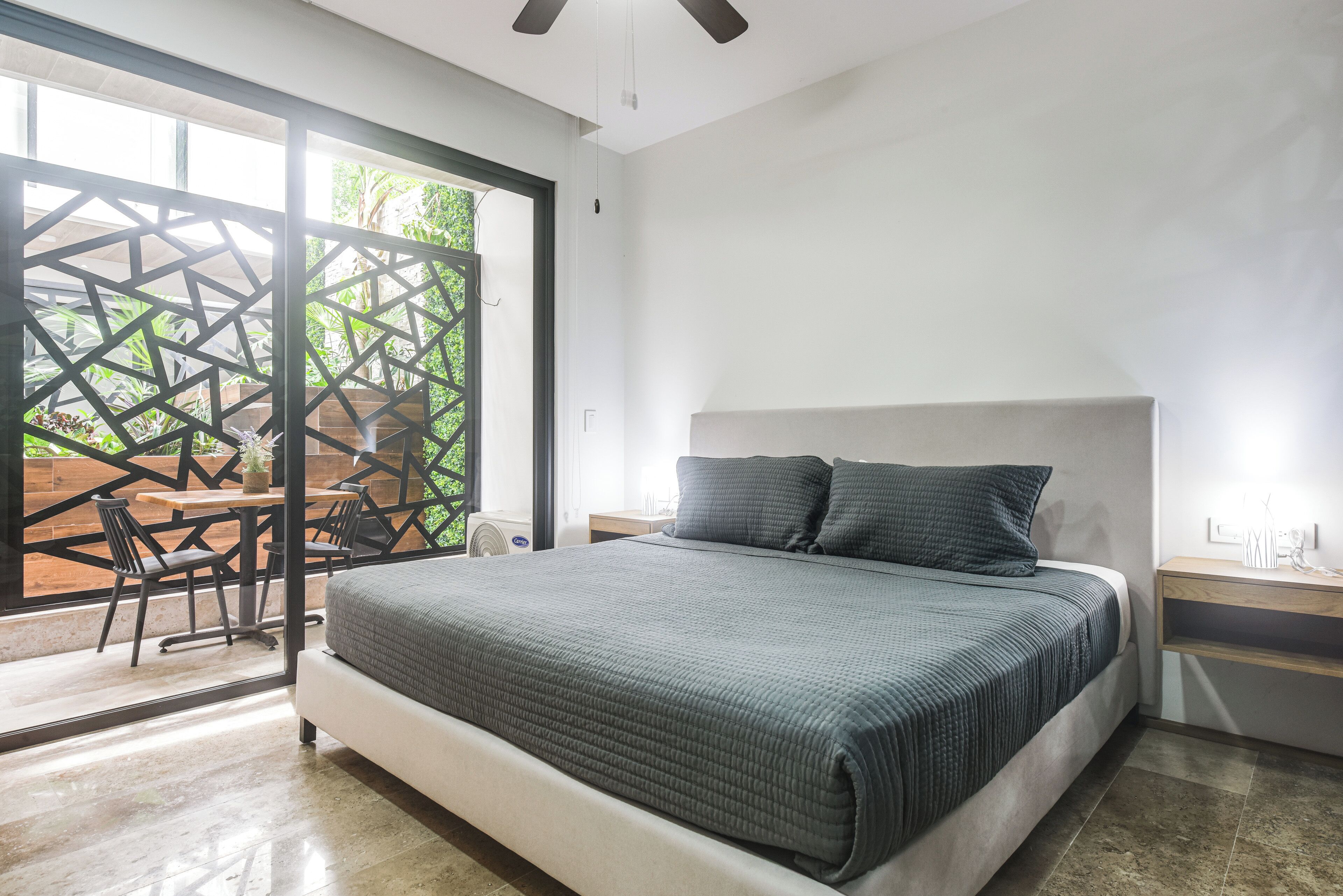 Une chambre moderne avec un grand lit, une table de chevet en bois, un ventilateur de plafond et une porte-fenêtre coulissante donnant sur une terrasse extérieure.