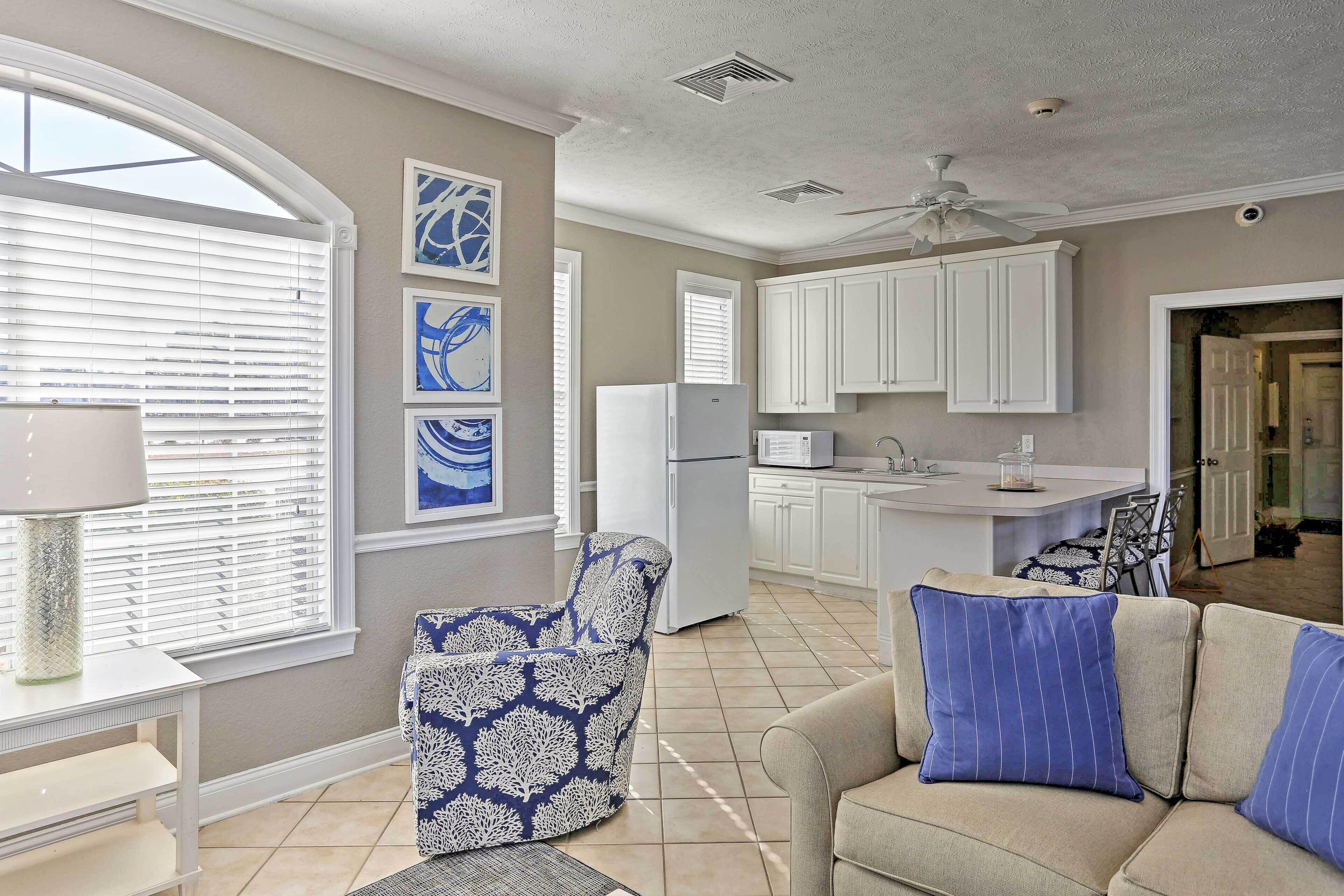 A modern kitchen with white cabinets, a refrigerator, and a microwave. There is a dining area with a table and chairs, and a living room with a sofa and cushions.