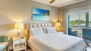 A bedroom with a large bed, two bedside tables with lamps, a green chair, a view of the outdoors, and a ceiling fan.