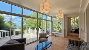 Um quarto espaçoso com janelas amplas, piso de madeira, mesa de vidro e corrimão branco na área externa.