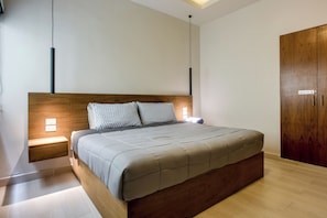 Ein modernes Schlafzimmer mit einem Holzkopfende, einem Bett mit grauer Bettdecke, Nachttischlampen und einem Kleiderschrank.