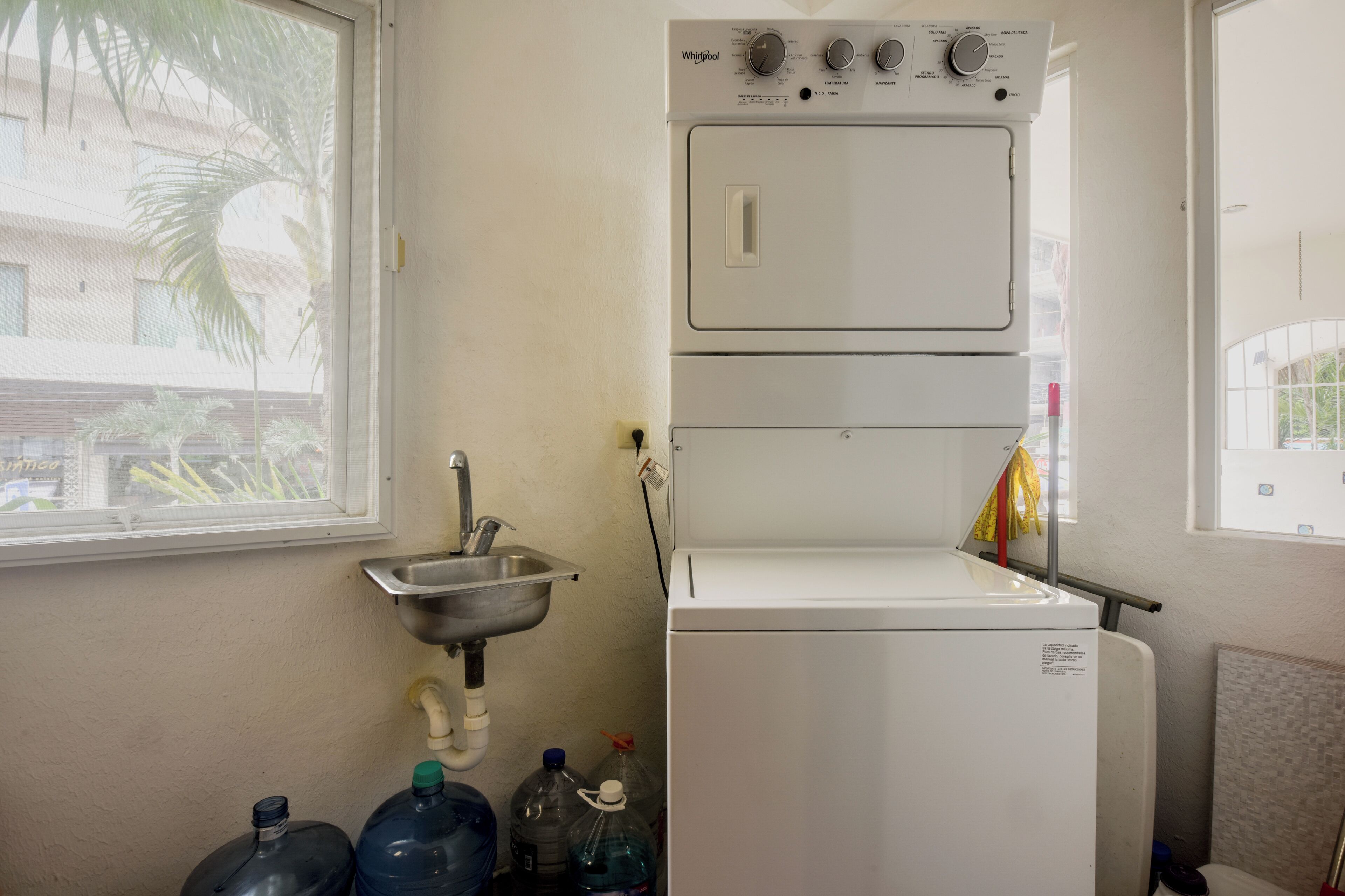 A compact laundry room with a stacked washer and dryer, a sink, and large water jugs.
