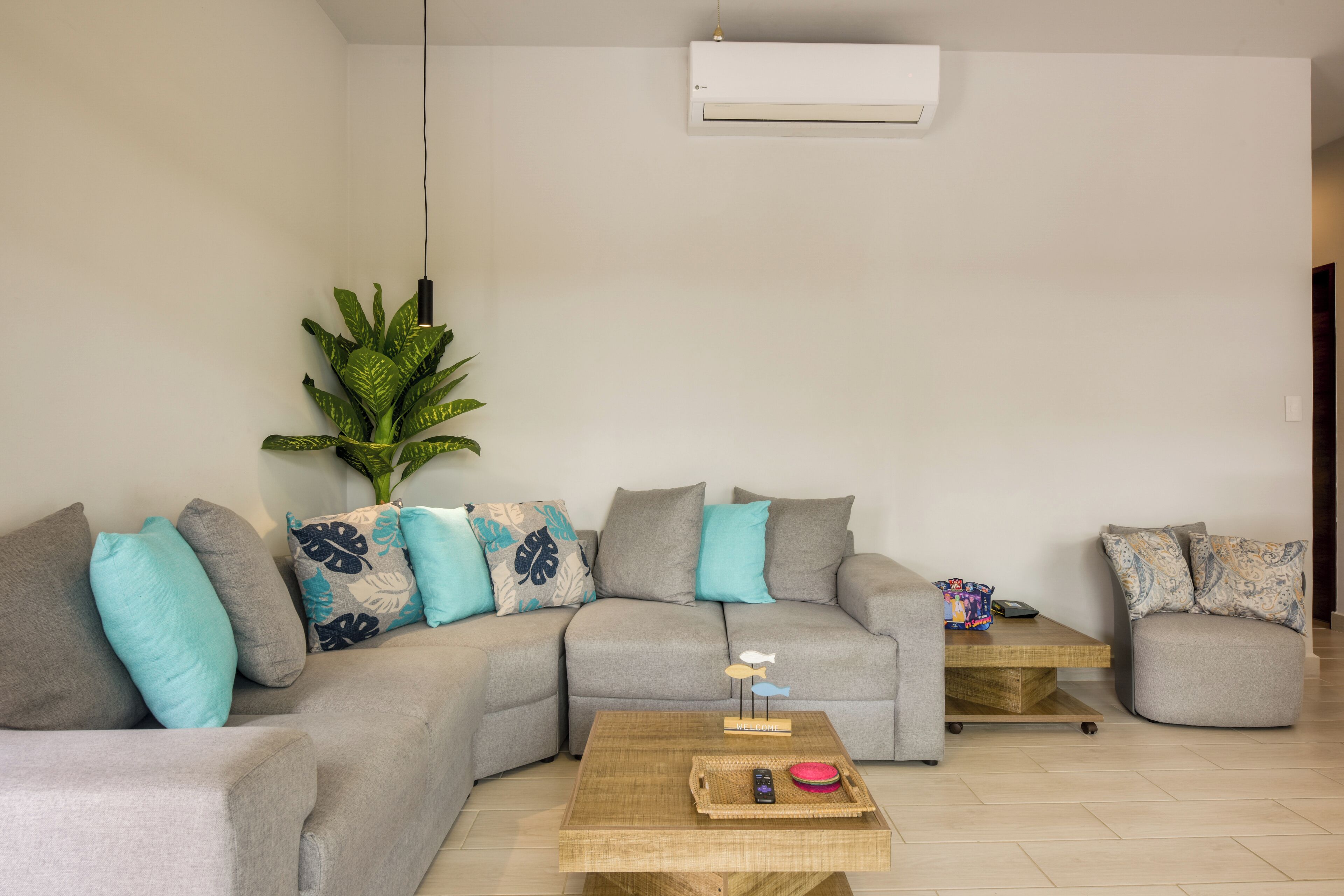 Una sala de estar con un sofá seccional, una mesa de centro, una planta en maceta y un aire acondicionado instalado en la pared.