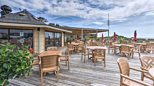 Une terrasse extérieure avec des tables et des chaises en bois, un espace salon couvert et une vue sur la mer.
