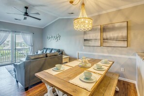 A dining area with a wooden table set for four, a grey sofa, a ceiling fan, and a chandelier. - Waterfront Condo w/ Views & Pool in Little River (Little River)