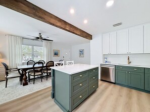Una cocina moderna con armarios blancos, una isla verde con cajones, lavavajillas integrado y un comedor con mesa y sillas.