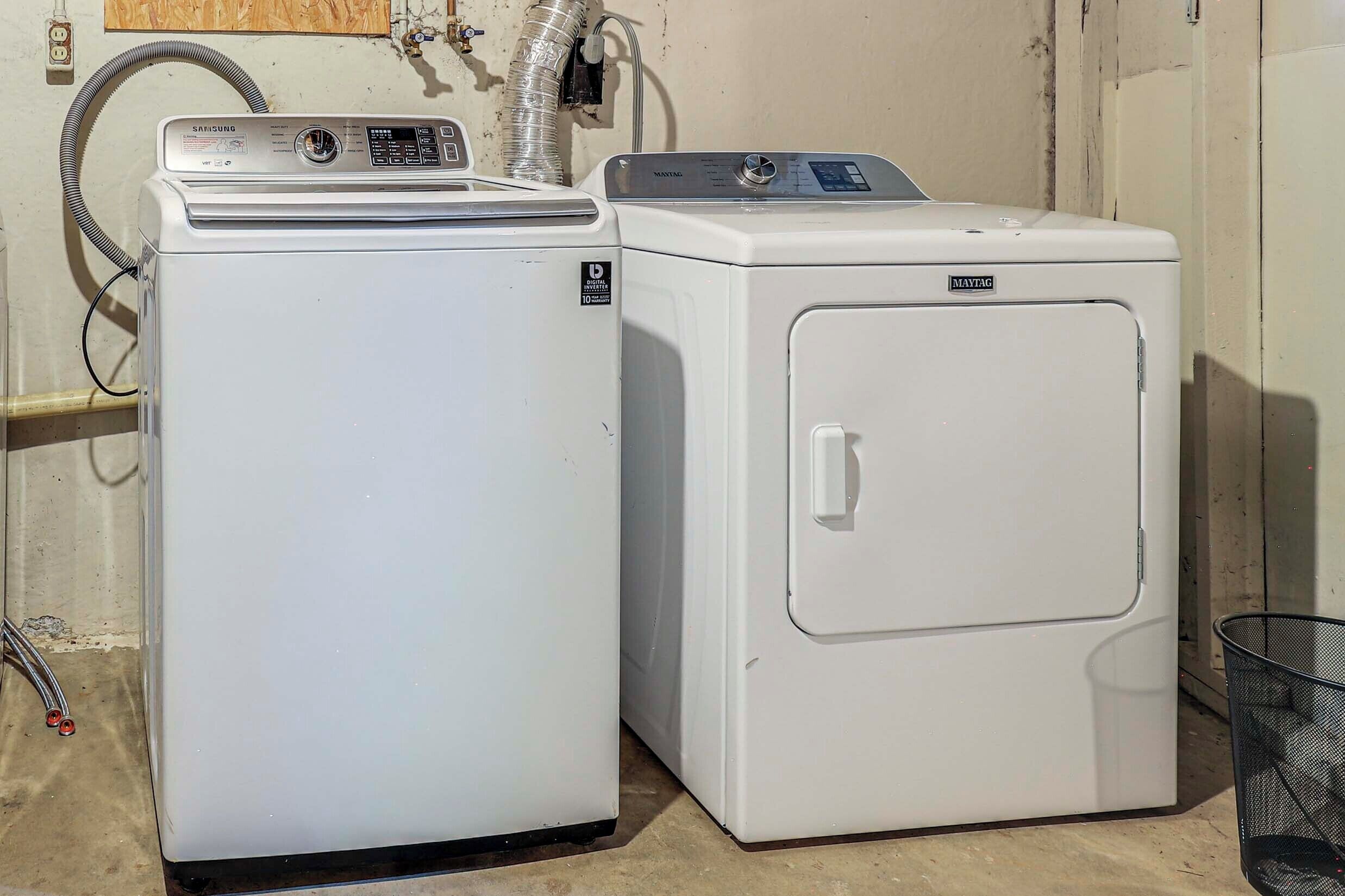 Two white washing machines, one with a control panel and the other with a front door, in a room with a concrete floor and a wall-mounted electrical outlet.