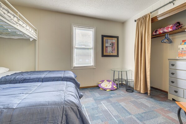 A bedroom with two beds, a window with blinds, a framed picture on the wall, a coat rack with hangers, and a dresser with drawers.