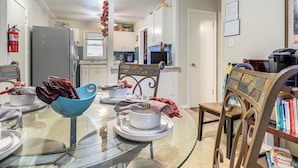 Una cocina con una mesa de comedor, sillas y un tazón azul. Hay un refrigerador, una cafetera y un extintor.