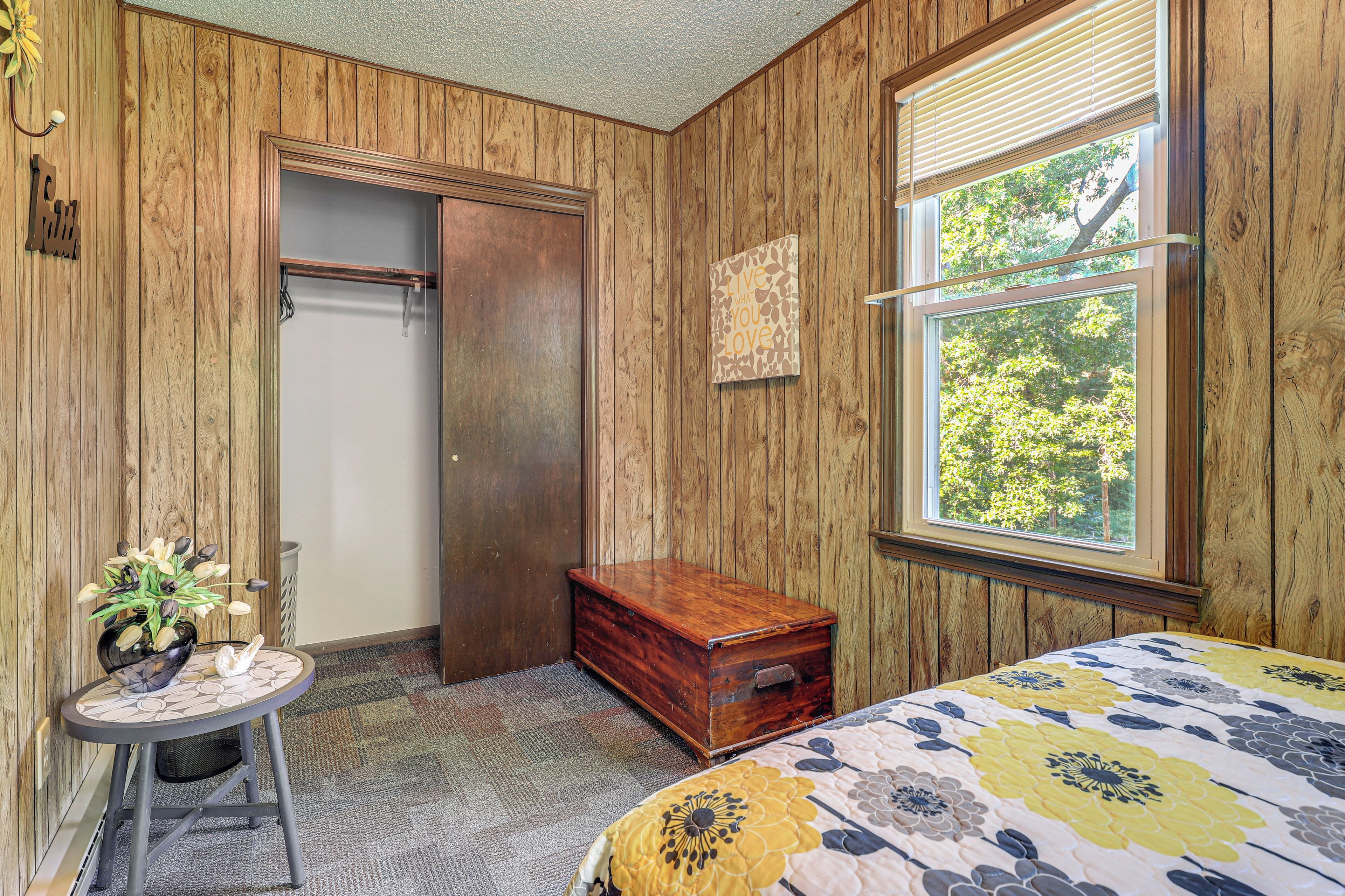 A room with wooden paneling, a bed with a floral bedspread, a small wooden chest, a table with a vase, and a window with a view of trees.