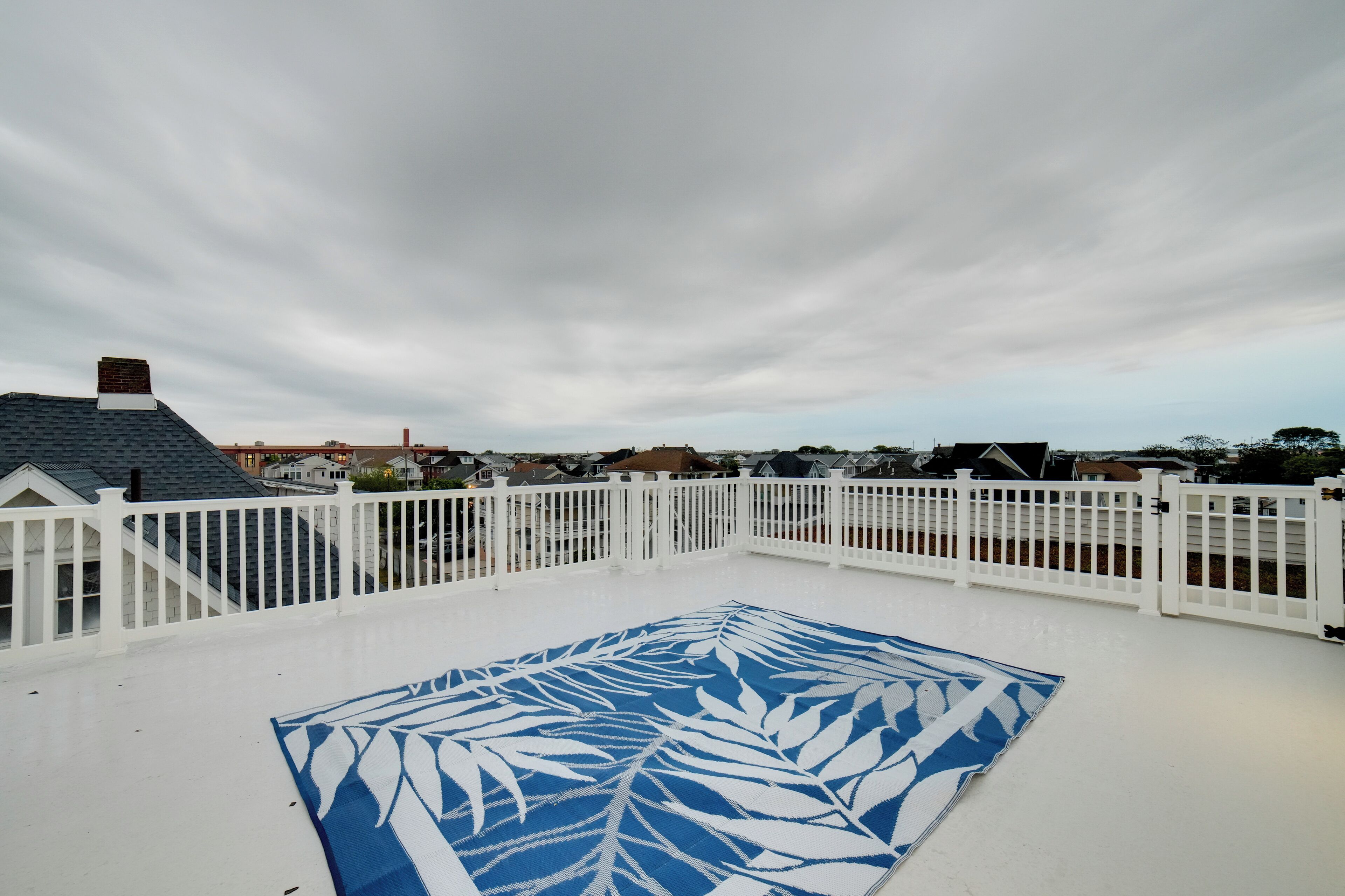 Eine Dachterrasse mit weißem Geländer, einer blauen, gemusterten Matte und Blick auf Häuser im Hintergrund.