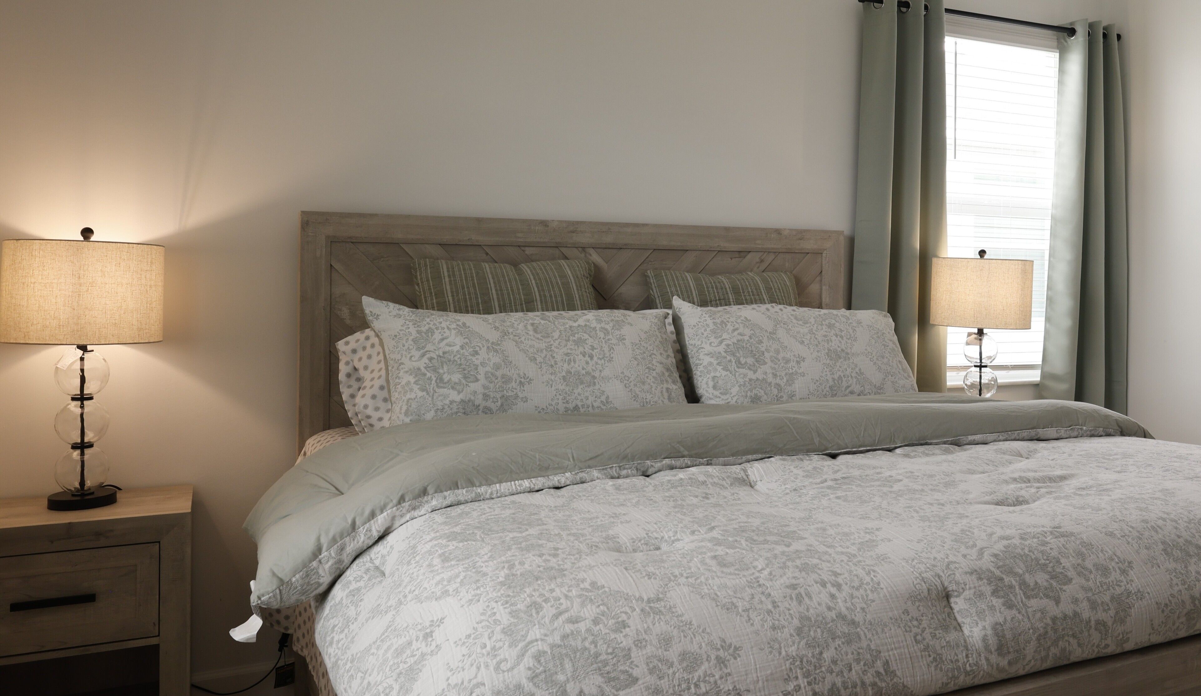 Quarto com cama, duas luminárias de cabeceira, mesa de cabeceira e janela com cortinas.