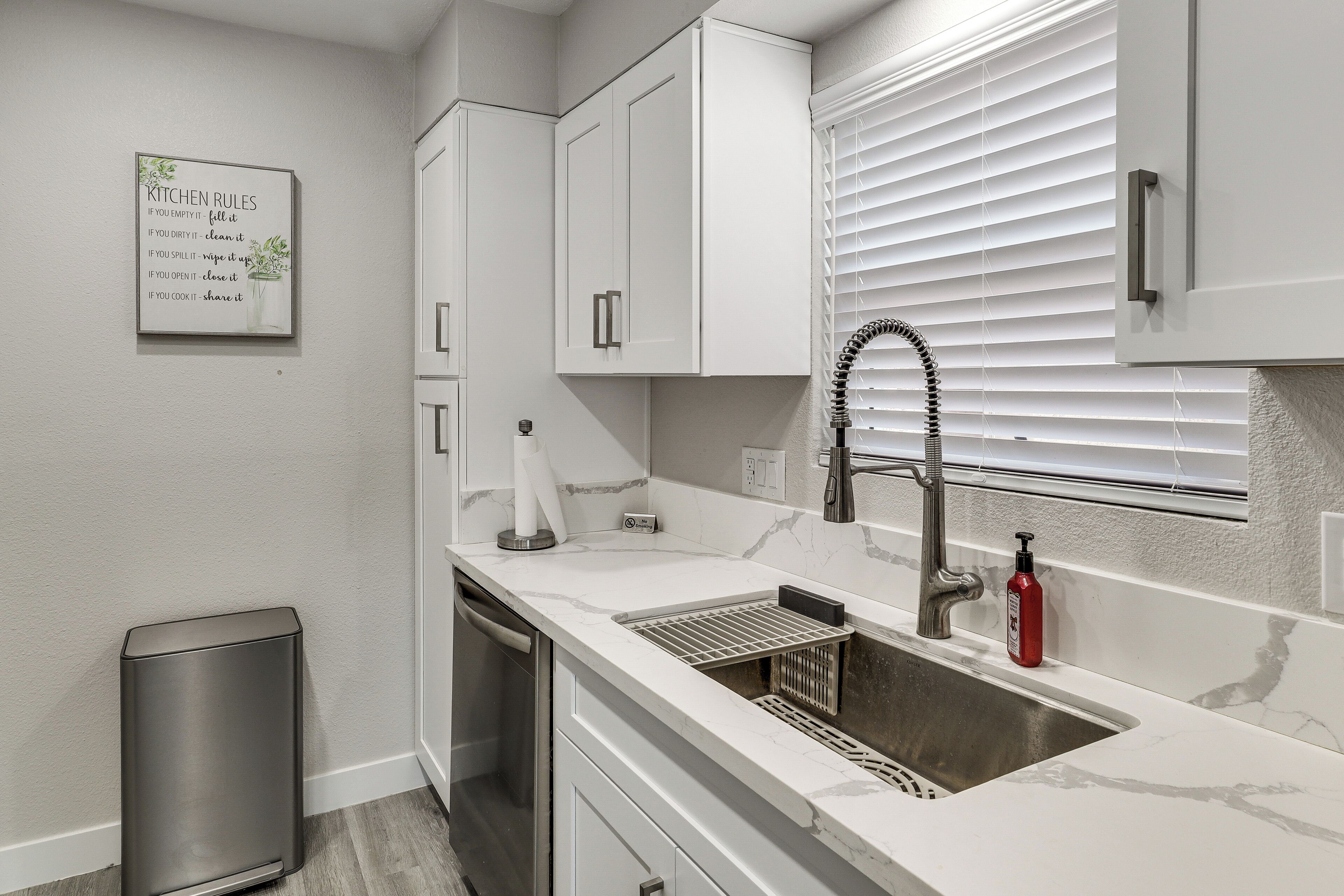 A kitchen with white cabinets, a stainless steel sink, a dish rack, and a trash can.